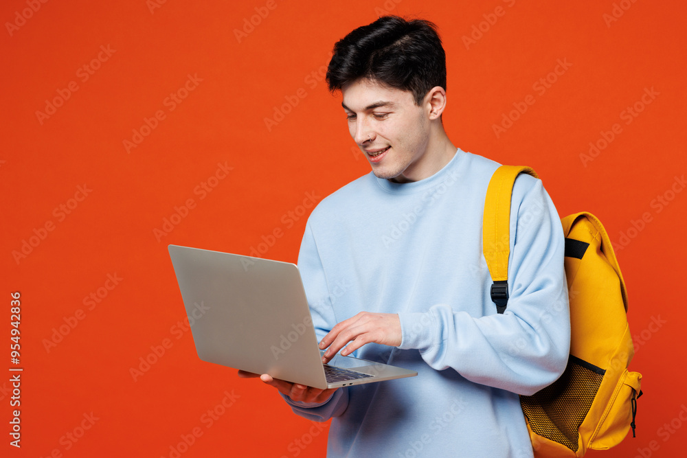 Young smiling happy fun man IT student he wearing blue casual clothes backpack bag hold use work on laptop pc computer isolated on plain red orange background. High school university college concept. Young smiling happy fun man IT student he wearing blue casual clothes backpack bag hold use work on laptop pc computer isolated on plain red orange background. High school university college concept.