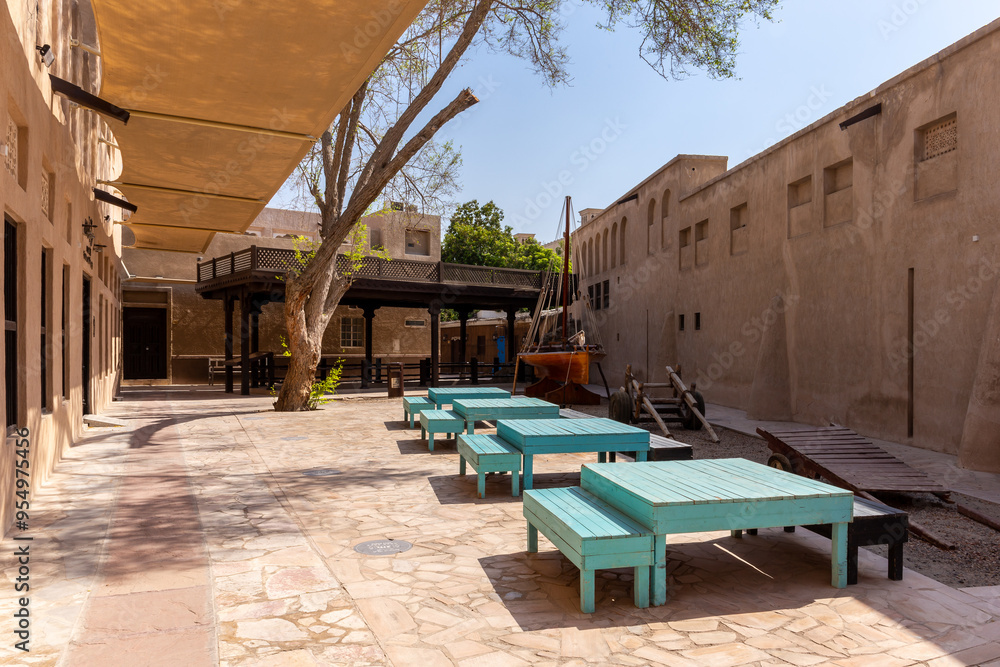 Fototapeta premium Al Fahidi Historical District in Dubai, stone street with traditional Arabic style buildings, turquoise tables, wooden traditional boat (dhow) and sunshades above, Deira.