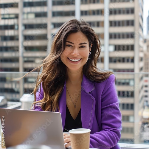 Young woman in purple blazer smiles while working on a laptop at a café with a city skyline view in the background, enjoying her coffee