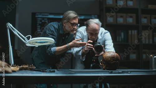 Two archaeologists photograph fossil remains of ancient human skull with professional camera on tripod, have discussion. Scientists or historians work in modern archaeological lab. Archaeology concept photo