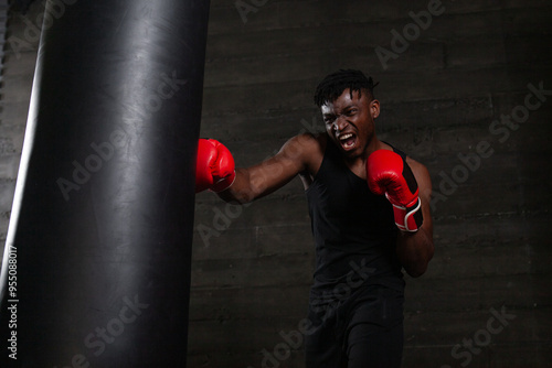 An African American boxer with a furious expression on his face delivers a powerful punch to a punching bag in a dark gym, demonstrating his strength and determination to defeat his opponent in the