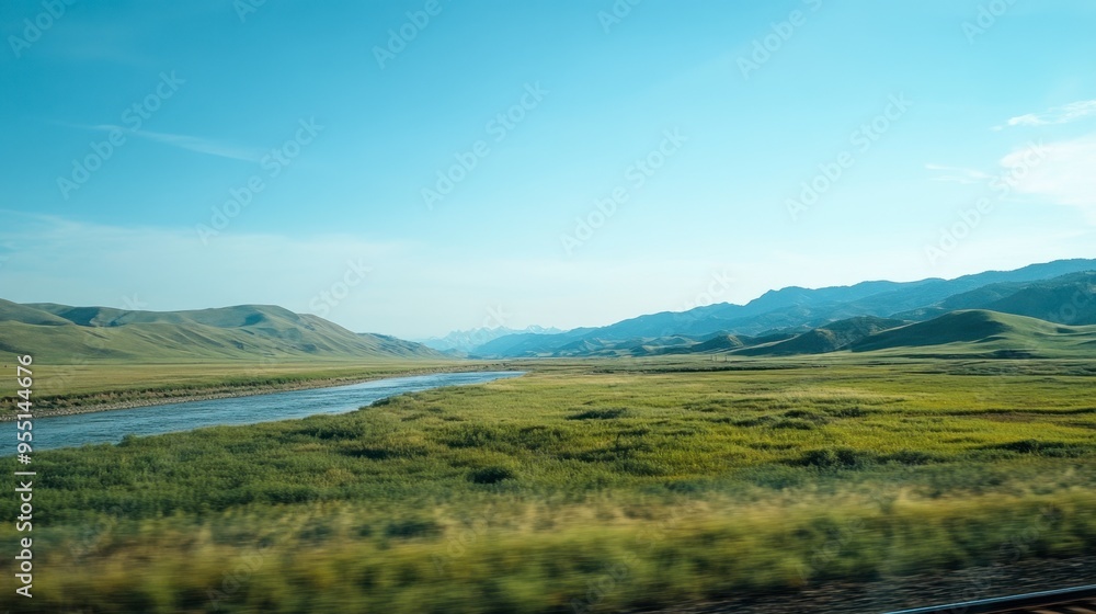 Fototapeta premium A scenic view from a train window, capturing rolling green hills, distant mountains, and a river running parallel to the tracks, under a clear blue sky.