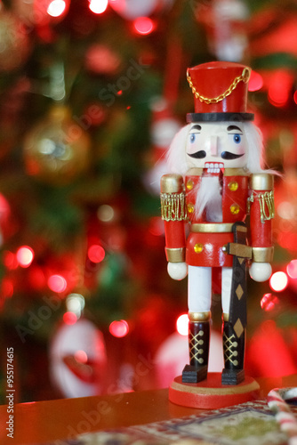 A red and gold nutcracker soldier figurine stands on a surface in front of a blurred, festive Christmas tree with glowing lights and ornaments, creating a warm holiday ambiance.