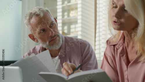 Tired pensive Caucasian couple family grandparents senior mature female woman elderly retired male man talk about business working with documents utility papers bills bank payment laptop sofa at home