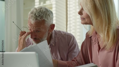 Tired pensive Caucasian couple family grandparents senior mature female woman elderly retired male man talk about business working with documents utility papers bills bank payment laptop sofa at home