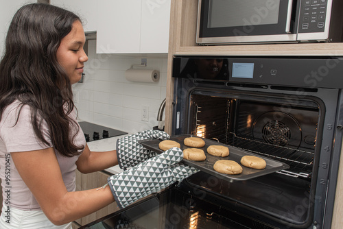Young teenager wearing mittens putting raw cookies on the hot oven.