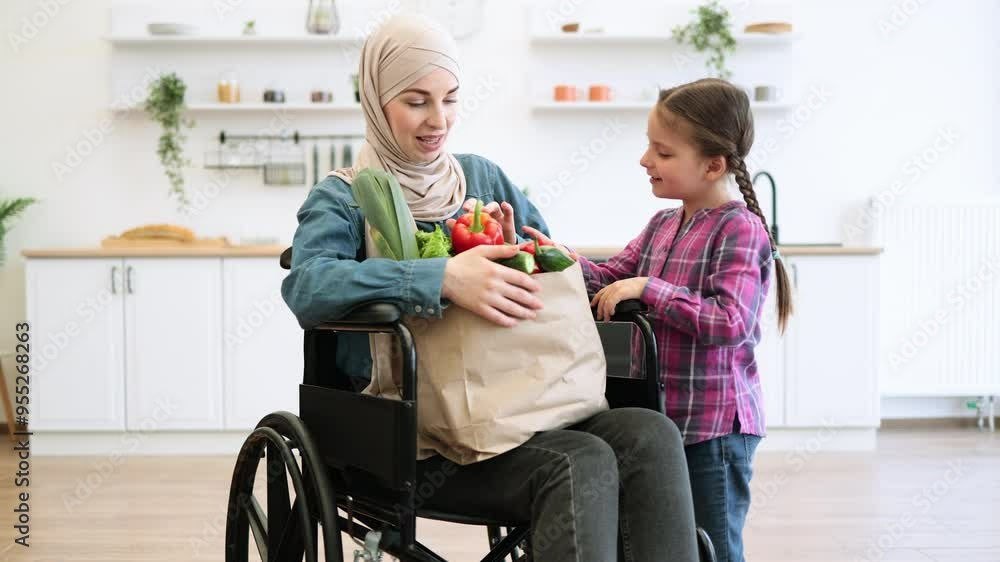 Cheerful woman in wheelchair and young daughter smiling while holding grocery bag filled with vegetables in bright kitchen. Concept of family support, inclusivity, and healthy living