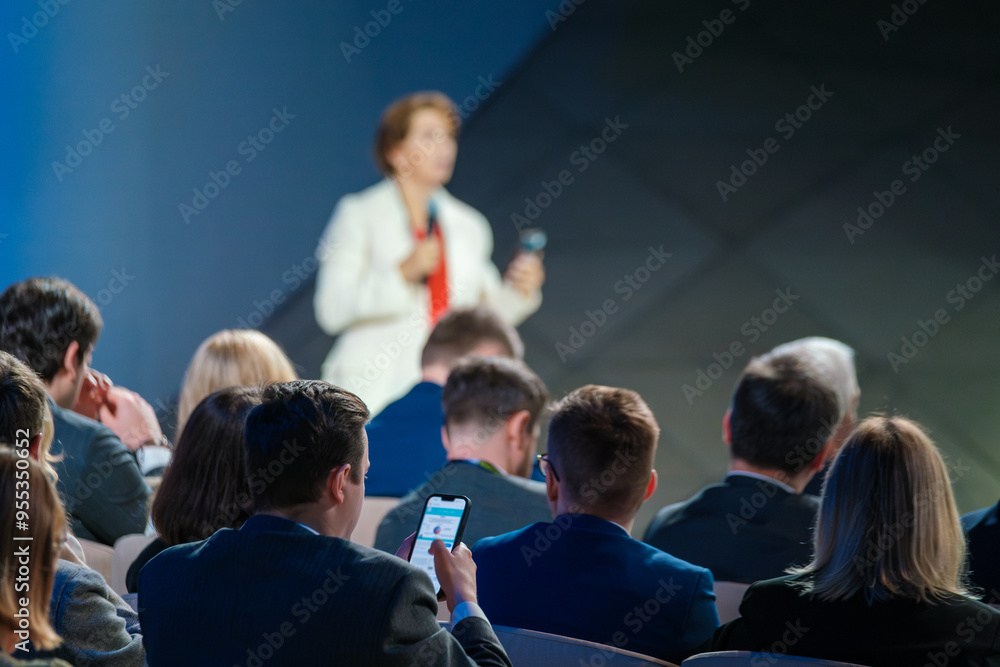 Obraz premium Audience members listening to a speaker during a business conference. Professionally dressed individuals engaged in a formal presentation.