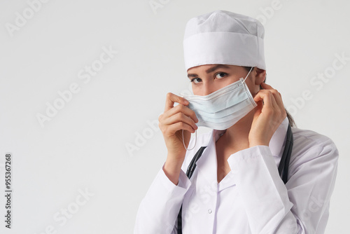 Portrait of young female doctor putting on protective medical face mask on white background, copy space