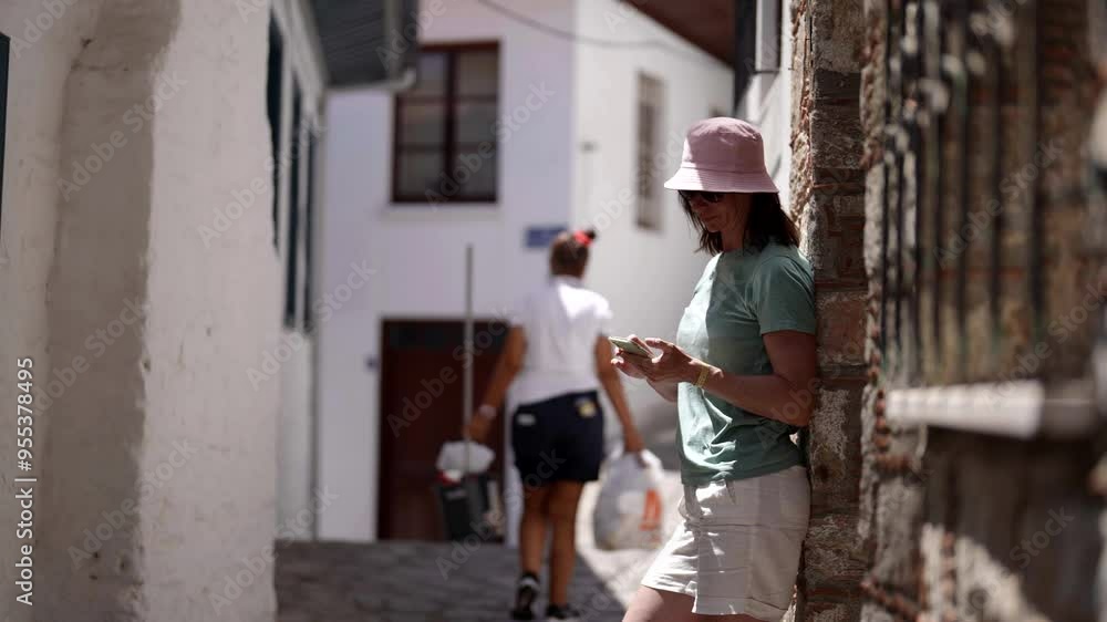 A slender woman in a pink panama hat and a green swimsuit is relaxing on an ancient street. She is focused on her phone, enjoying the peace and warmth. A slender woman in a pink panama hat and a green swimsuit is relaxing on an ancient street. She is focused on her phone, enjoying the peace and warmth.