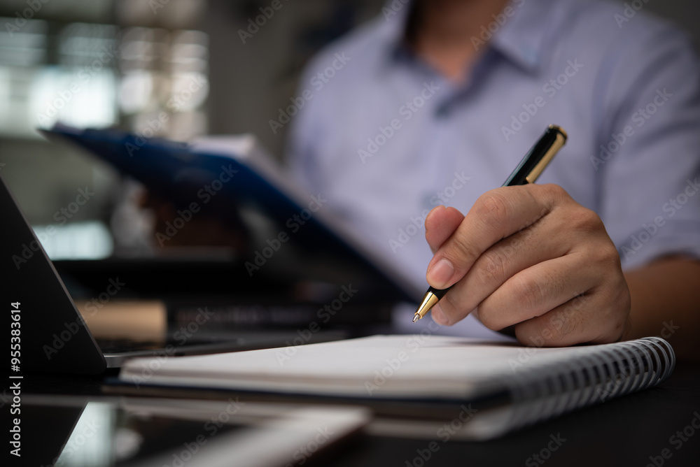 Business professional taking notes in a notebook while reviewing documents at a desk planning organization and business strategy development. Represents business analysis. Business professional taking notes in a notebook while reviewing documents at a desk planning organization and business strategy development. Represents business analysis.