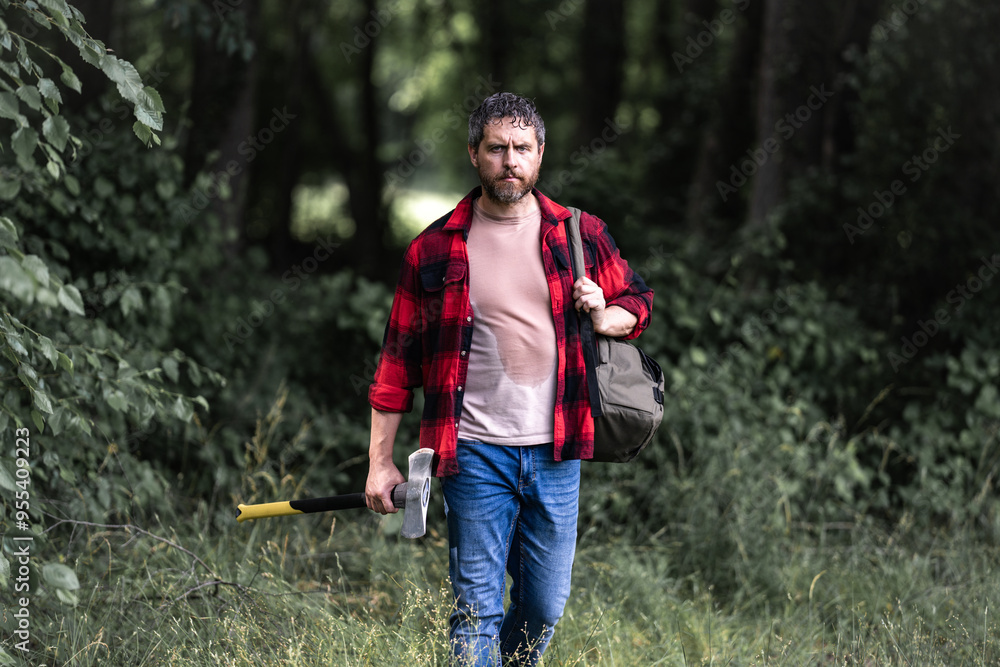 The Lumberjack holds axe with serious face on the forest. A lumberjack with a large ax examines the tree before felling. Serious lumberjack. Confident young bearded man holding a big axe outdoor. The Lumberjack holds axe with serious face on the forest. A lumberjack with a large ax examines the tree before felling. Serious lumberjack. Confident young bearded man holding a big axe outdoor.