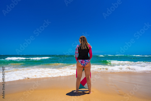 Beautiful blonde woman in long sleeve swimsuit standing with multicolored bodyboard and looking at big waves on sunny beach Bordeira praia, Algarve, Portugal. Back view. Water sports in summertime. photo