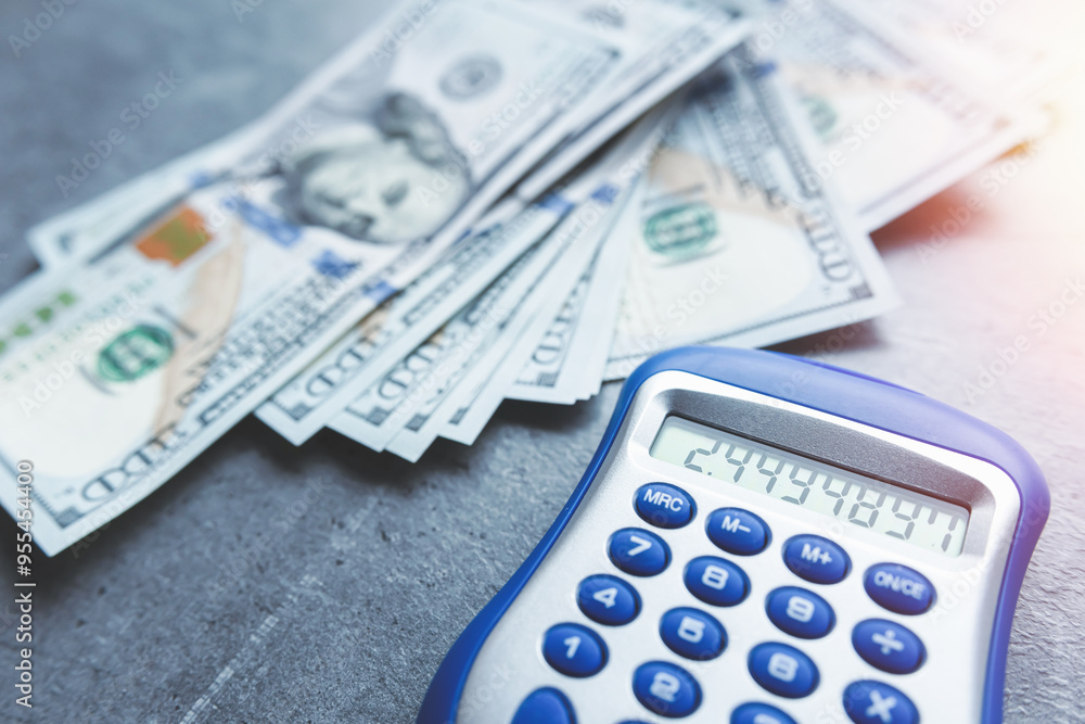 Many dollar bills and calculator on grey desk. Investment research data economy. Many dollar bills and calculator on grey desk. Investment research data economy.