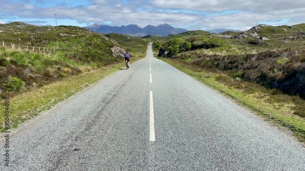 Cyclist riding along straight road through landscape to mountains beyond Cyclist riding along straight road through landscape to mountains beyond