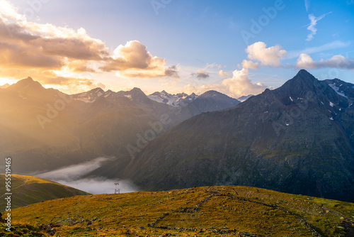 The sun rises over the majestic Otztal Alps, casting a golden glow on the peaks and valleys. Clouds linger softly above the landscape, creating a stunning contrast at dawn in serene mountain region photo