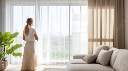 A woman gazes out a large window in a modern living room filled with natural light during the day, surrounded by soft furnishings and plants