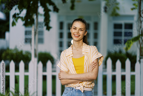 Smiling young woman standing confidently in front of a white picket fence She wears a yellow top and a light striped shirt, radiating positivity against a lush green background