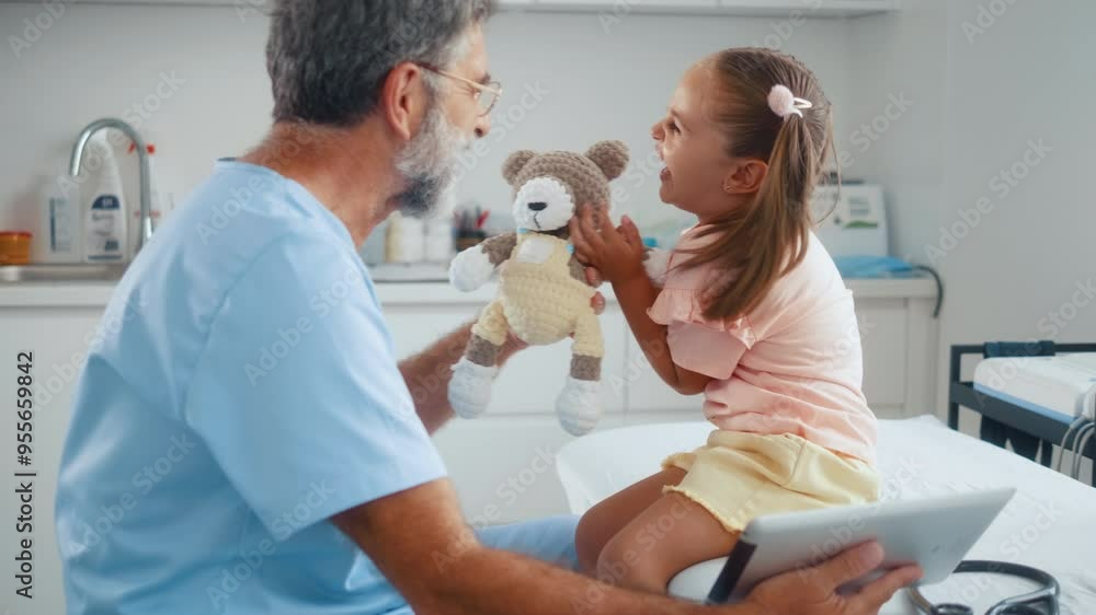 Senior male medical worker holds a teddy bear and makes a little girl patient laugh during a medical examination in the hospital Senior male medical worker holds a teddy bear and makes a little girl patient laugh during a medical examination in the hospital