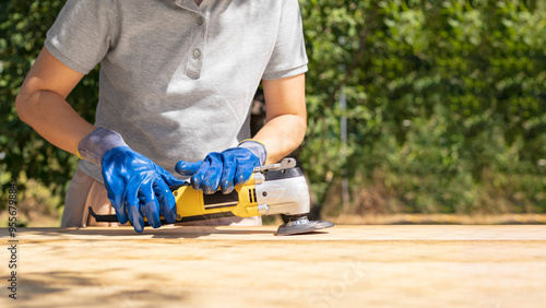 Carpenter Woman Wearing Protective Gloves Using Power Sander For Sanding Wooden Plank Outdoors. Concept Of Reconstruction And Restoration Of Wooden