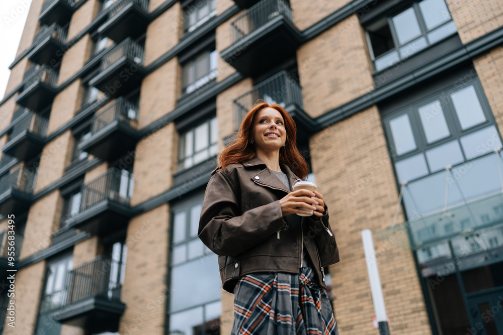 Low-angle view of cheerful redhead woman savoring coffee break in urban environment, holding cup and sipping drink with smile, exuding joy and relaxation, standing on background of modern building. Low-angle view of cheerful redhead woman savoring coffee break in urban environment, holding cup and sipping drink with smile, exuding joy and relaxation, standing on background of modern building.