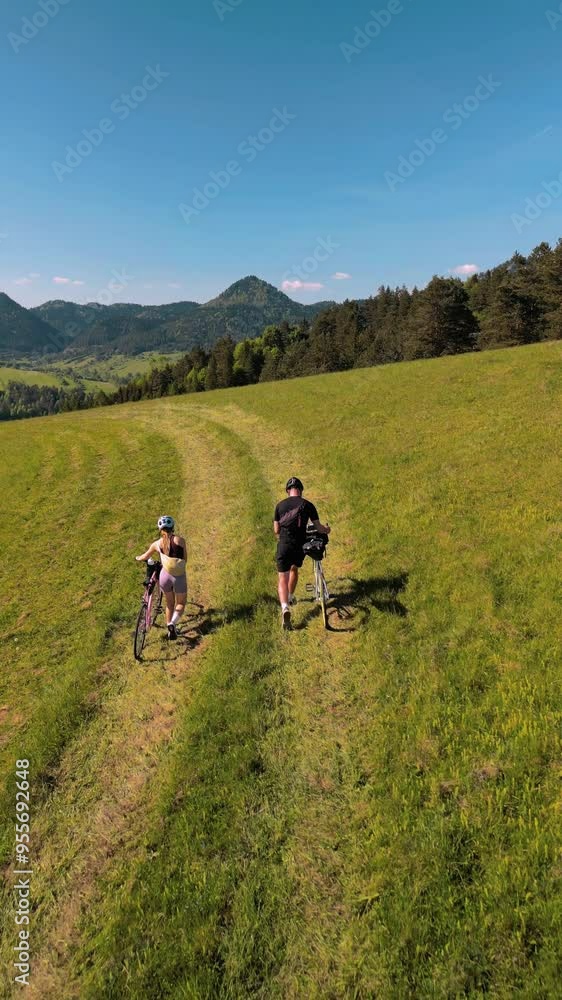 Trip to the mountains on bicycles. Two cyclists in the mountains. Two cyclists are pushing their bicycles. A man and a woman are riding their bicycles in a field with mountains in the background. Trip to the mountains on bicycles. Two cyclists in the mountains. Two cyclists are pushing their bicycles. A man and a woman are riding their bicycles in a field with mountains in the background.