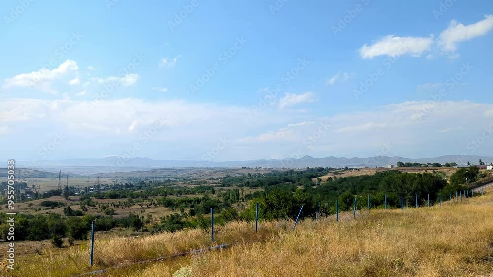 A panoramic view of the countryside. Poles with wires, dry grass and several trees are visible in the foreground. In the background are rolling fields, small villages and mountains on the horizon. A panoramic view of the countryside. Poles with wires, dry grass and several trees are visible in the foreground. In the background are rolling fields, small villages and mountains on the horizon.