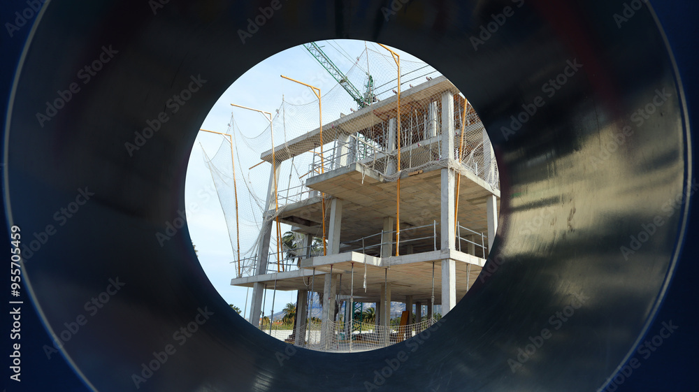 A construction site reveals a multi-story building under development, framed by a large pipe. Workers are busy on the structure amid clear blue skies. A construction site reveals a multi-story building under development, framed by a large pipe. Workers are busy on the structure amid clear blue skies.