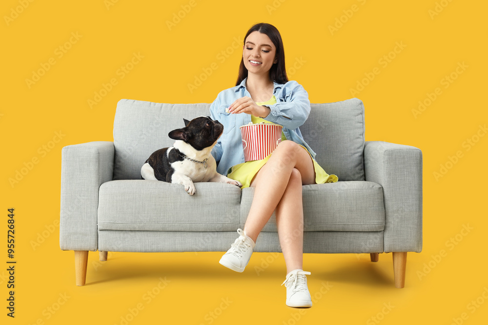 Young woman with her French bulldog eating popcorn on sofa against yellow background Young woman with her French bulldog eating popcorn on sofa against yellow background