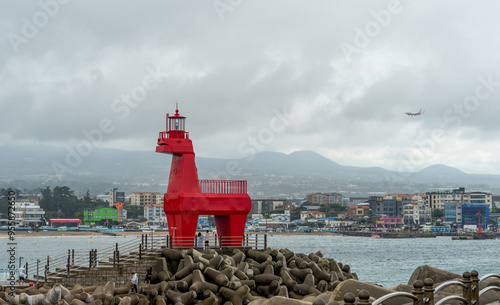 Jeju-si, Jeju-do, South Korea - August 16, 2021: Tourists are under a red lighthouse of horse shape on breakwater with tetrapods against houses and Ihoteu Beach photo