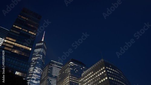 A cluster of modern office buildings and skyscrapers with brightly lit windows, including the One World Trade Center, contrasts against the deep blue night sky in New York City's financial district.