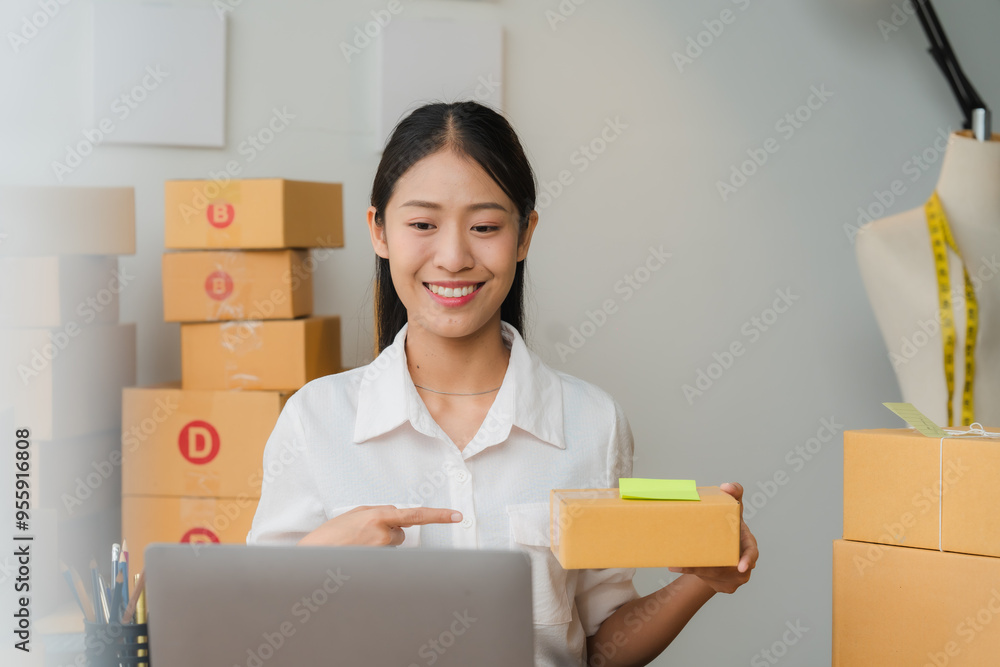 Smiling Entrepreneur: A young, Asian businesswoman confidently shows off a product in her home office. Surrounded by cardboard boxes, she radiates positivity and entrepreneurial spirit. The image evok Smiling Entrepreneur: A young, Asian businesswoman confidently shows off a product in her home office. Surrounded by cardboard boxes, she radiates positivity and entrepreneurial spirit. The image evok