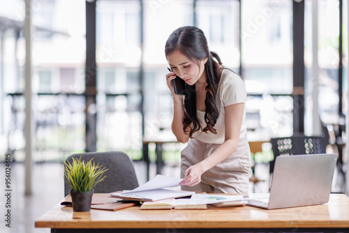 Smiling asian female hold cell at ear talk look at laptop screen. Busy young asian lady advisor lawyer give phone consultation from workplace discuss electronic document details