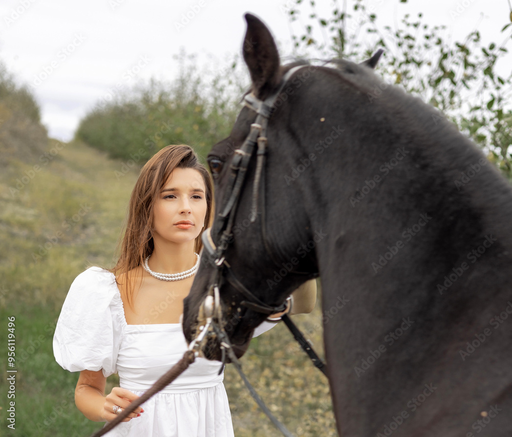 Obraz premium beautiful,delicate woman in white dress posing with black scary dark horse. girl and animal in orchard in garden.female is trying to pet the horse.