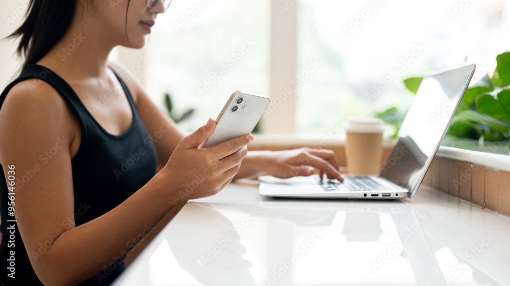 A woman working remotely from a coffee shop, holding her smartphone while working on her laptop. A woman working remotely from a coffee shop, holding her smartphone while working on her laptop.
