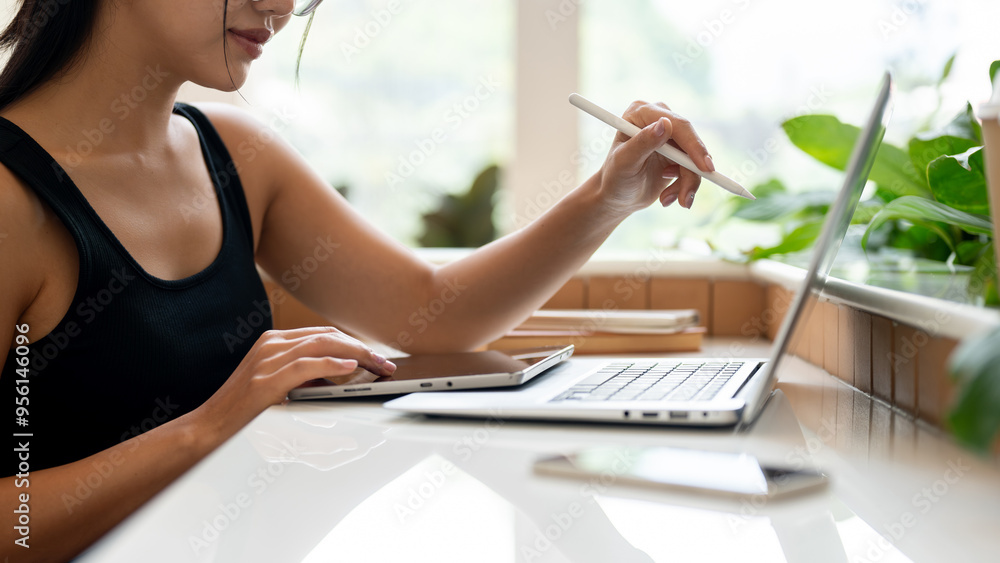 Obraz premium A beautiful Asian woman working remotely from a coffee shop, working on her laptop and tablet.
