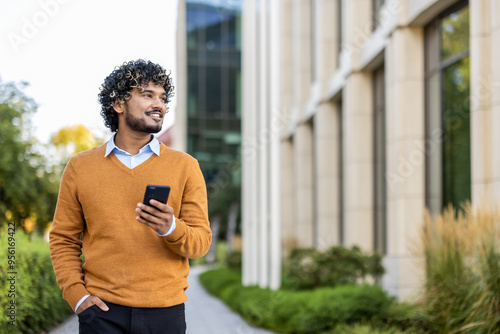 Hispanic businessman in orange sweater holding phone while walking outside office building. Smiling confidently and looking forward, showcasing modern urban work environment , professional lifestyle. photo