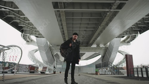 Young man in modern urbanistic recreation area for city dwellers in outskirts. Handsome hipster looking at camera, zooming shot, slow motion, contemporary lifestyle in town, infrastructure for sports