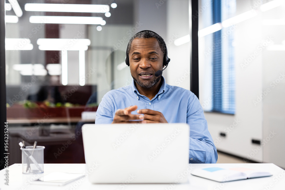 Confident businessman wearing headset participates in online meeting. Sitting at desk with laptop, engaging in professional communication. Focused on business discussions Confident businessman wearing headset participates in online meeting. Sitting at desk with laptop, engaging in professional communication. Focused on business discussions
