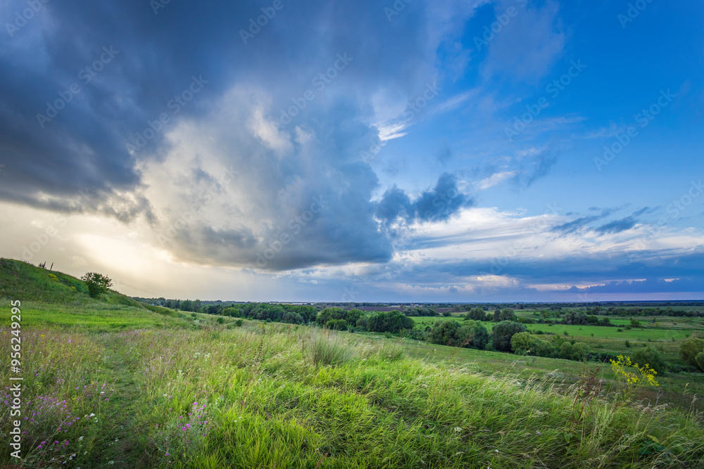 Vast green fields stretch beneath dramatic clouds, hinting at an approaching dusk that casts a soft glow over the serene valley landscape.