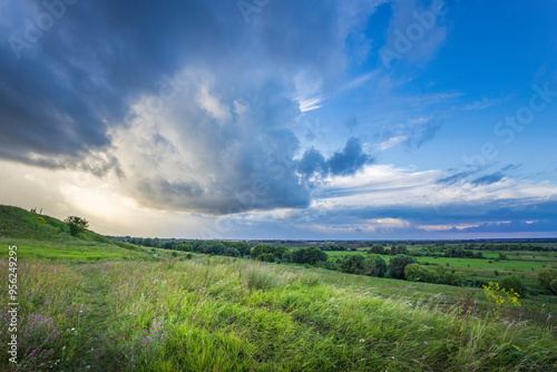 Obraz na płótnie Vast green fields stretch beneath dramatic clouds, hinting at an approaching dusk that casts a soft glow over the serene valley landscape. w salonie