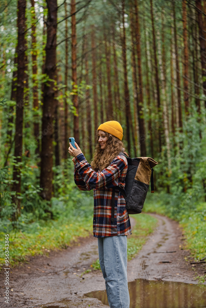 Curly woman hiker in yellow beanie and flannel shirt takes photo with smartphone while hiking through lush green forest Curly woman hiker in yellow beanie and flannel shirt takes photo with smartphone while hiking through lush green forest