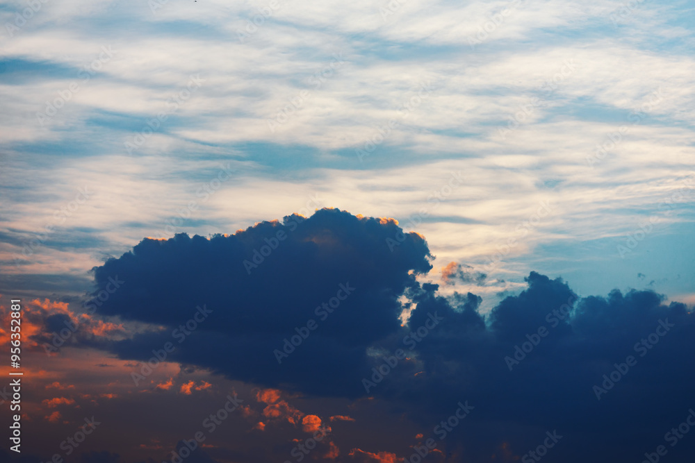 Fototapeta premium Cloudscape With Colored Clouds at Sunset. Large cloud in the sky with a bright orange sun peeking through it. The sky is mostly blue with a few clouds scattered throughout