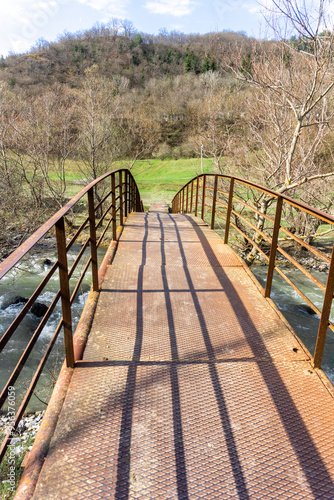 A rusty arched iron bridge with railings over the Tedzami River leads to a clearing with green grass. Mountains are in the background photo