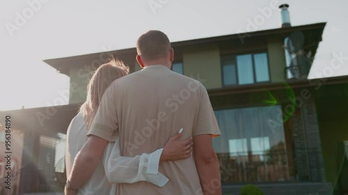 Back view of caucasian couple looking at new luxury house while standing at the yard. Young family dreaming about buying a new big house standing and hugging.