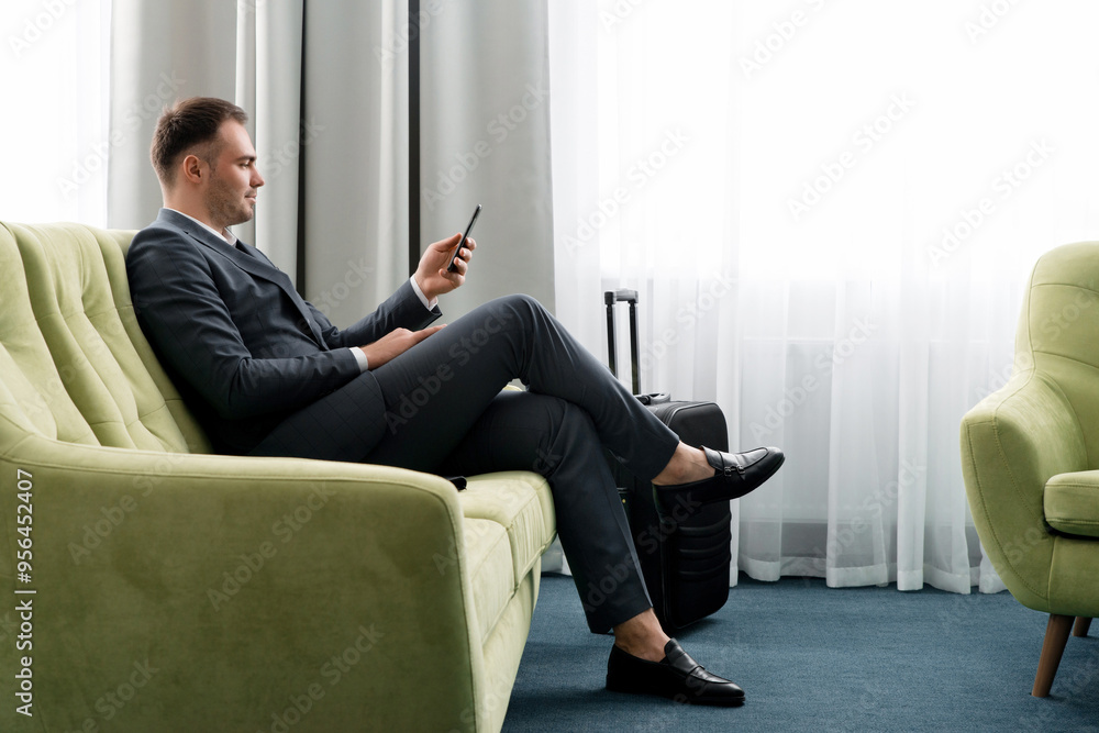 Cheerful businessman using phone sitting in hotel room with suitcase on business trip Cheerful businessman using phone sitting in hotel room with suitcase on business trip