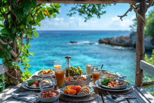 A panoramic view captures luxurious breakfast table set for two, adorned with an array of delectable dishes, against backdrop of a stunning tropical sea, creating a picturesque scene that epitomizes e photo