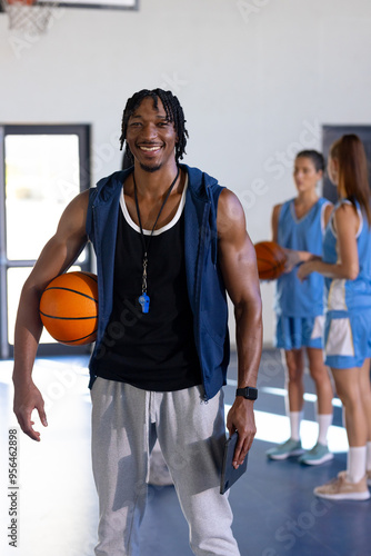 male african American basketball coach holding ball and whistle, smiling in gym with female players