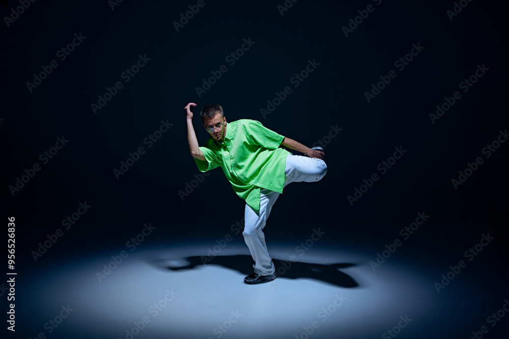 Young guy in jeans and green t-shirt dancing hip-hop with energy and rhythm against black studio background. Concept of street style, contemporary dance, entertainment