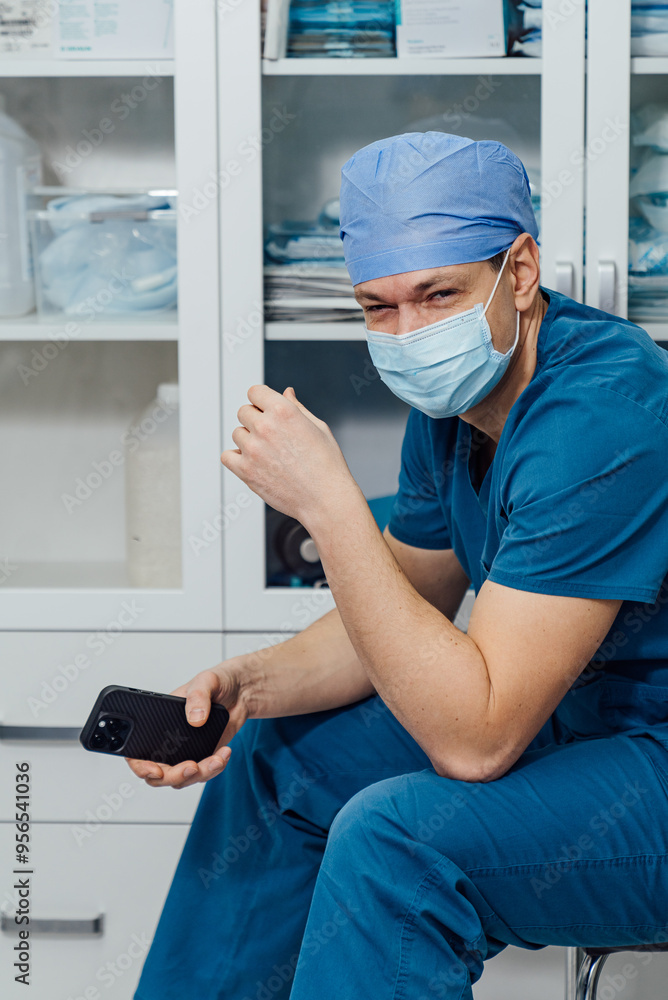 Surgeon in scrubs checking his phone while seated in a medical office. A surgeon wearing scrubs and a mask sits in a medical office Surgeon in scrubs checking his phone while seated in a medical office. A surgeon wearing scrubs and a mask sits in a medical office